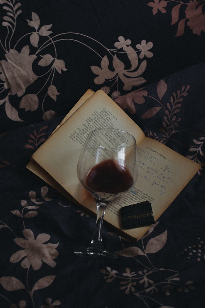 Open vintage book with a glass of red wine resting on floral fabric, evoking elegance and nostalgia.
