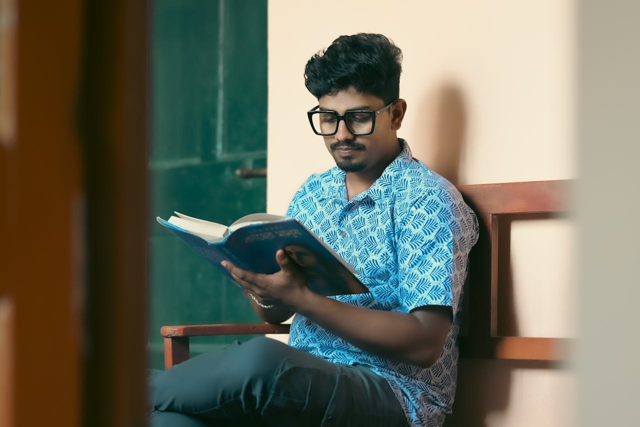 The Art of Drawing Readers In: Your attractive post title goes here A young man with glasses reading a book indoors, wearing a patterned shirt.