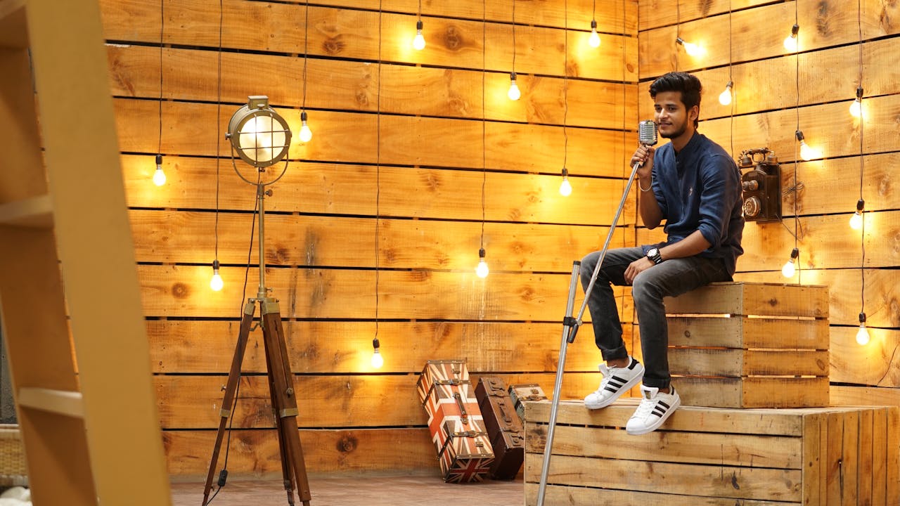 Young man singing on stacked crates with bright string lights and vintage decor.